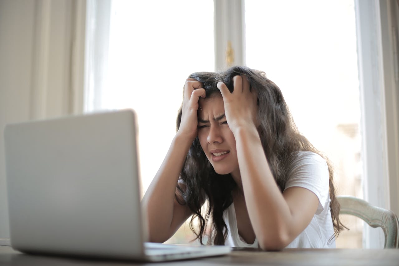 Frustrated young woman holding her head while sitting at a table with a laptop in front of a bright window.