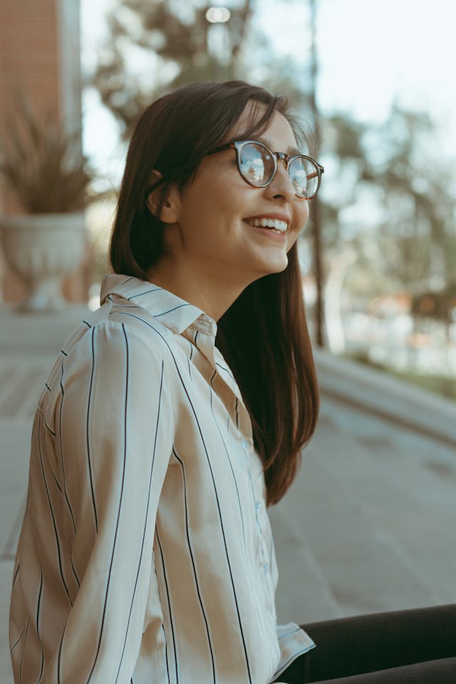 Smiling young woman with long dark hair and glasses wearing a white and black striped shirt sitting outdoors.